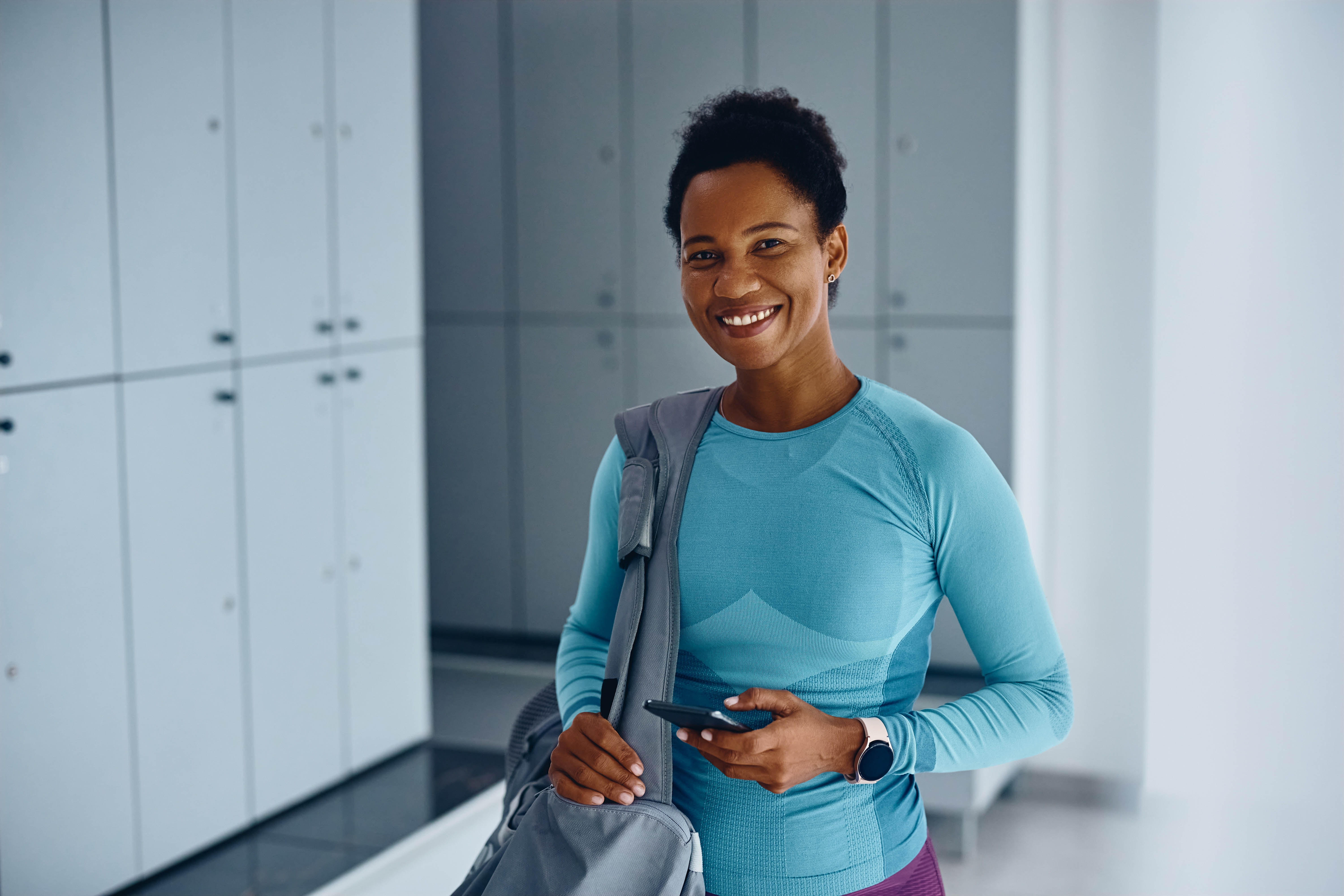 Middle aged woman standing and smiling, with her athletic gear in a bag over her shoulder, standing in a gym locker room